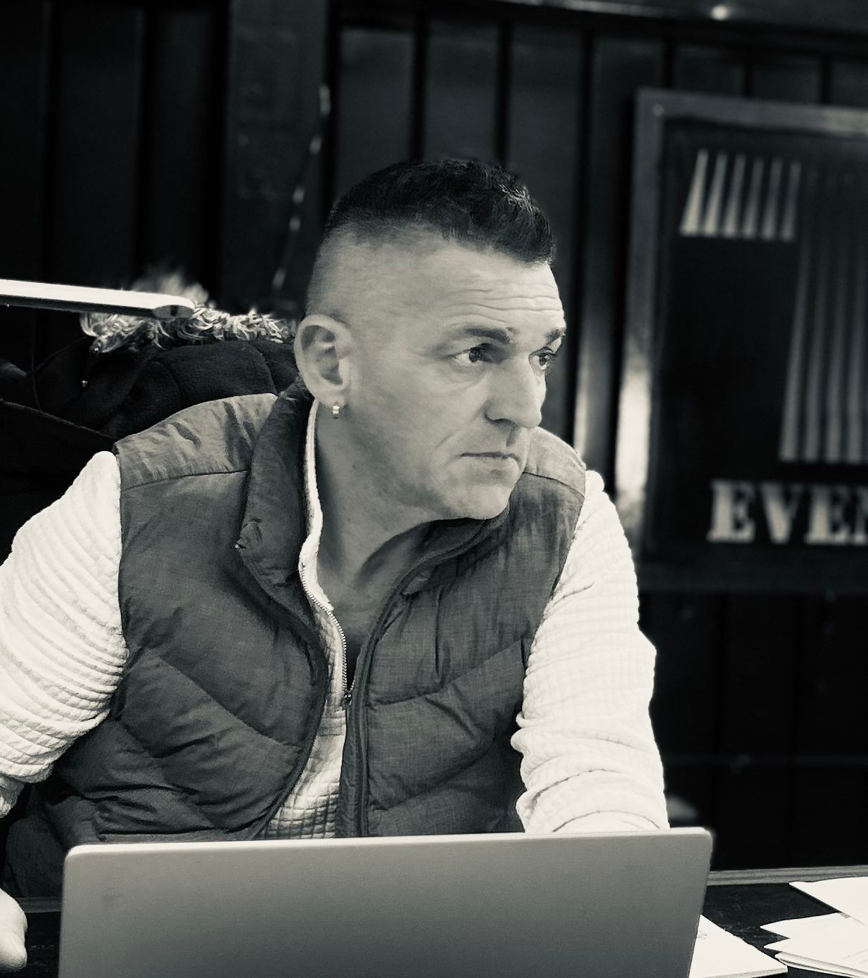 Man in a vest sitting at a table with a laptop, looking to the side.